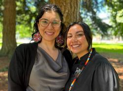 Two women stand among the trees in a park. They are close together and smile towards the camera.