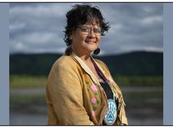 A woman wearing a buckskin jacket and a beaded medallion around her neck stands against a backdrop of a hilly range and cloud-filled sky. 