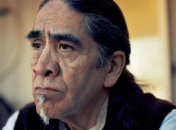 A man with heavy dark eyebrows and a grey patch of a goatee on his chin sits in a room and stares out of frame.