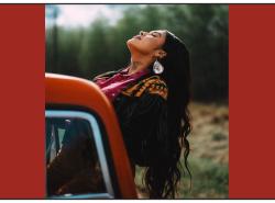 A young woman with long black hair leans backwards out the passenger side of a truck, closes her eyes and raises her face to the sun.