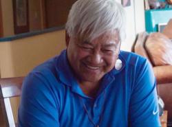 A man with grey hair smiles and looks downward. He is sitting in a kitchen chair.