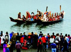 Two canoes filled with paddlers arrive at shore to a throng of people watching. Paddles are pointing up and dancers in regalia and masks stand on platforms built on the canoes' prow.