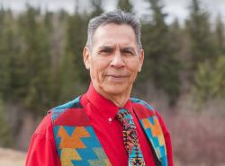 A man in a red shirt and multi-coloured vest and tie stands in front of a backdrop of trees.