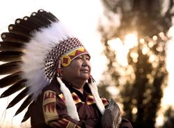 A man stands with his arms crossed across his chest. He has an eagle wing in his hand. He wears a shirt with geometric designs acress the front near his shoulder and down his arms. On his head is a feathered headdress. The same designs as on his shirt is beaded on the headband of the headdress.