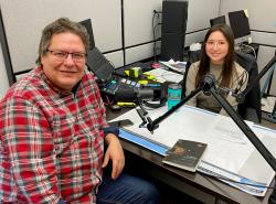 A man sits in a radio station booth with a young woman. Each has a radio microphone in from of them and notes on the desk top between them.